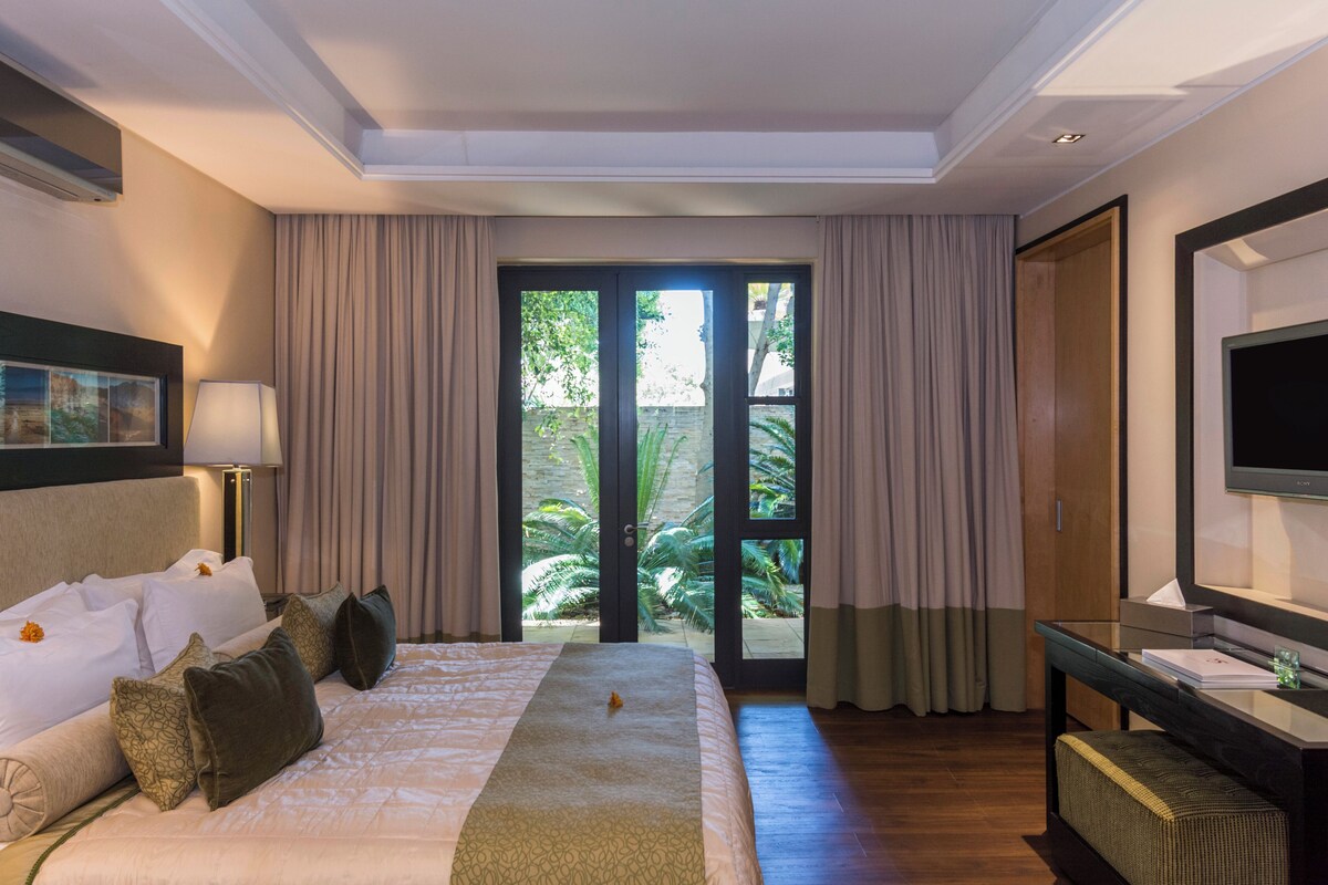 A hotel room with a large bed, a desk, a television, and a sliding glass door to a garden.
