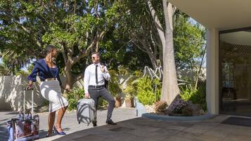 A man and a woman walking outside a building with a suitcase, surrounded by greenery and a clear sky.