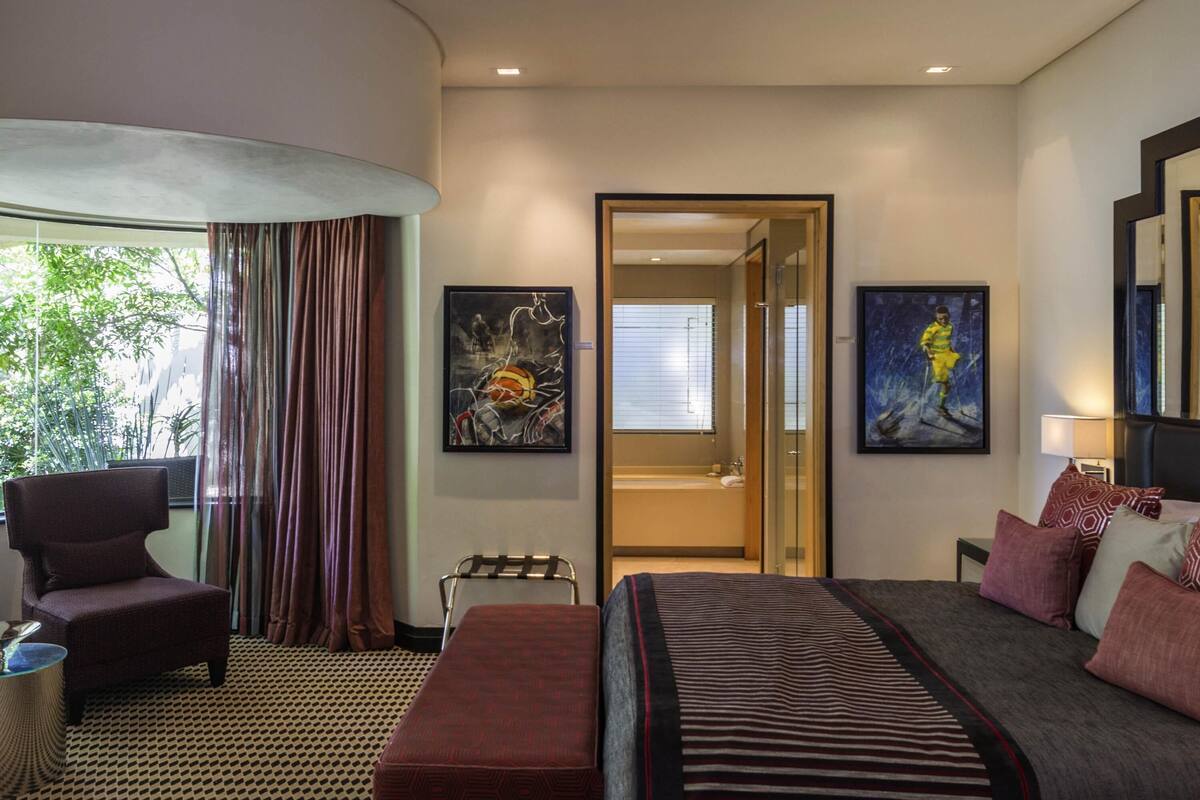 A hotel room with a bed, a chair, a small table, a bench, and artwork on the walls.