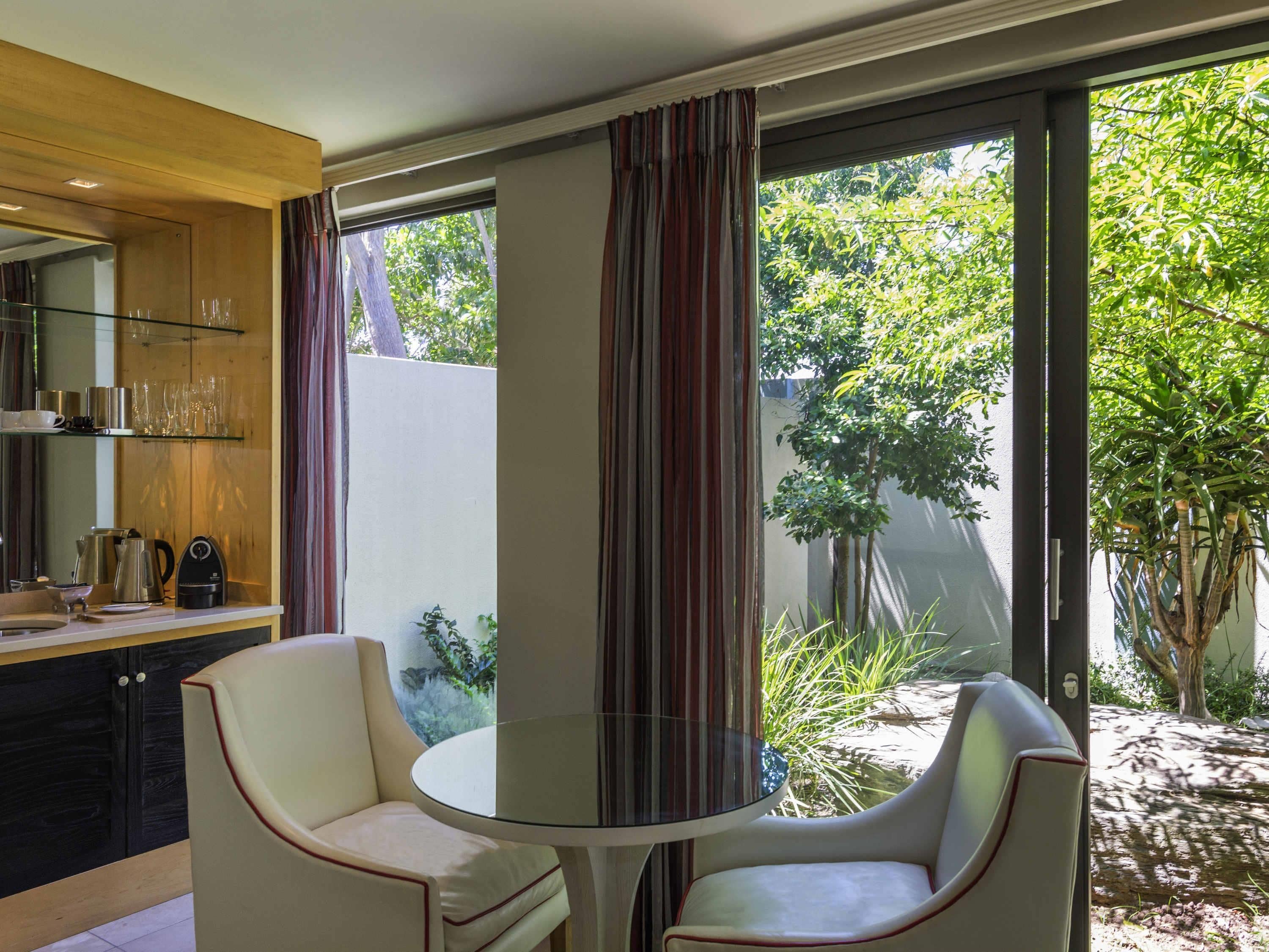A modern room with a glass table and two chairs, a kitchen area with a coffee maker, and large windows with curtains.