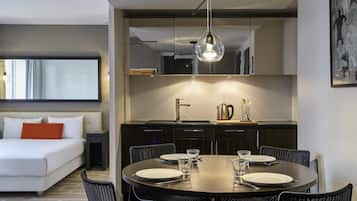 A modern hotel room with a bed, a dining table set for two, a kitchen area, and a large window.