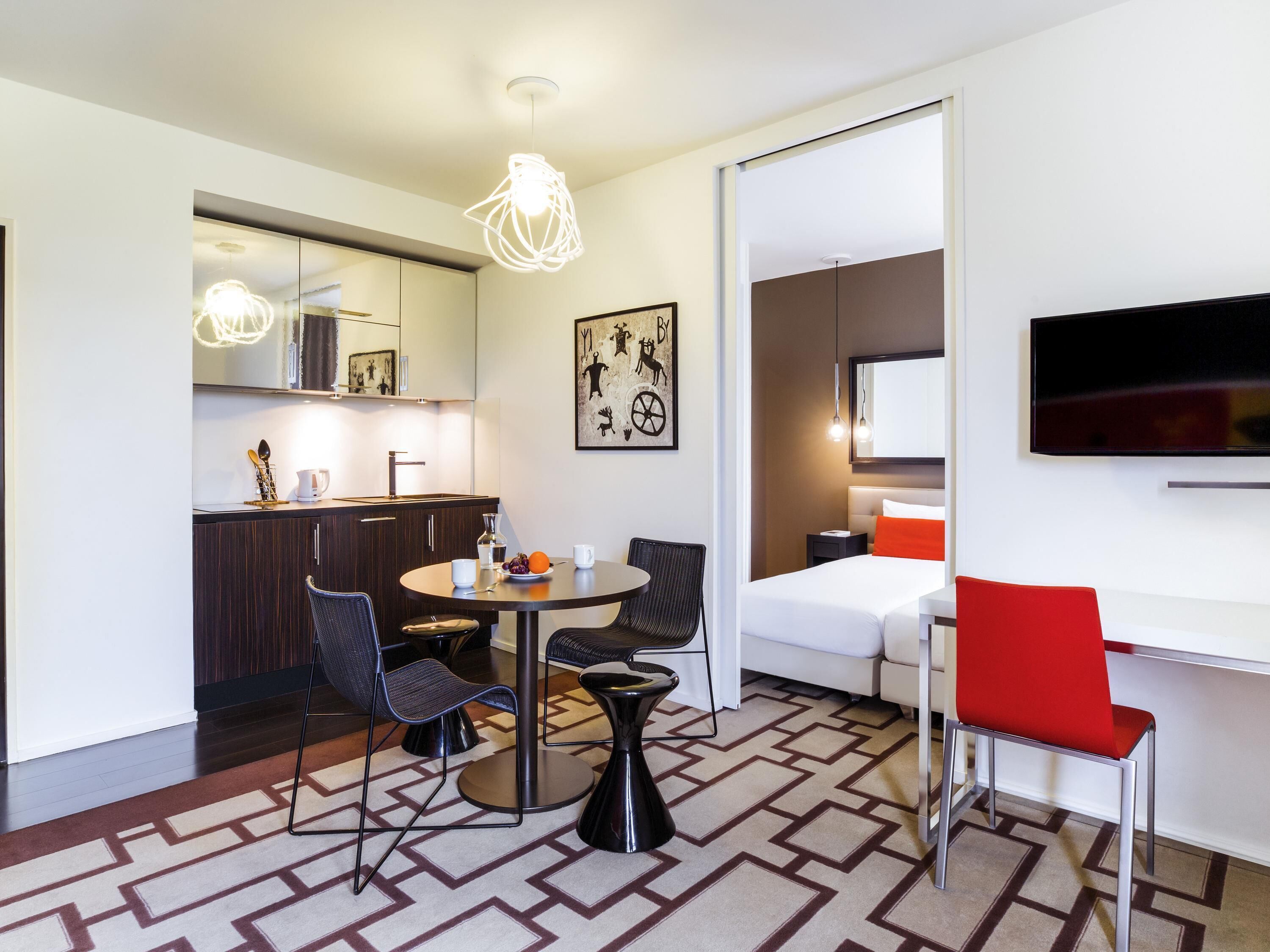 A modern hotel room with a dining area, kitchen, and a TV mounted on the wall.