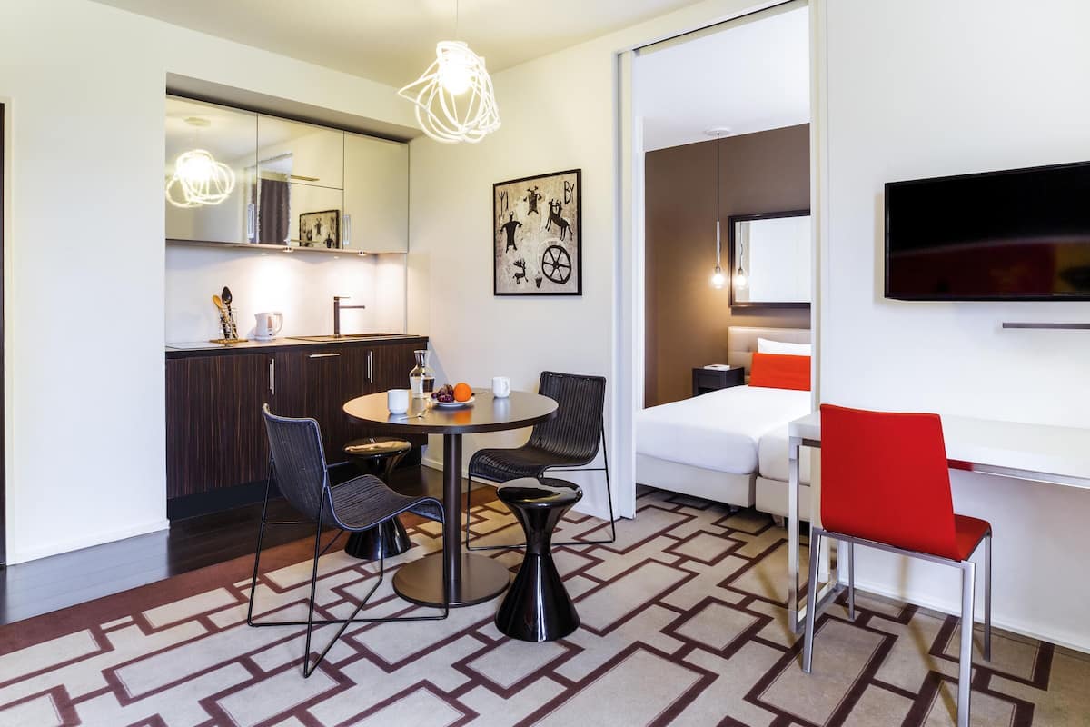 A modern hotel room with a dining area, kitchen, and a TV mounted on the wall.