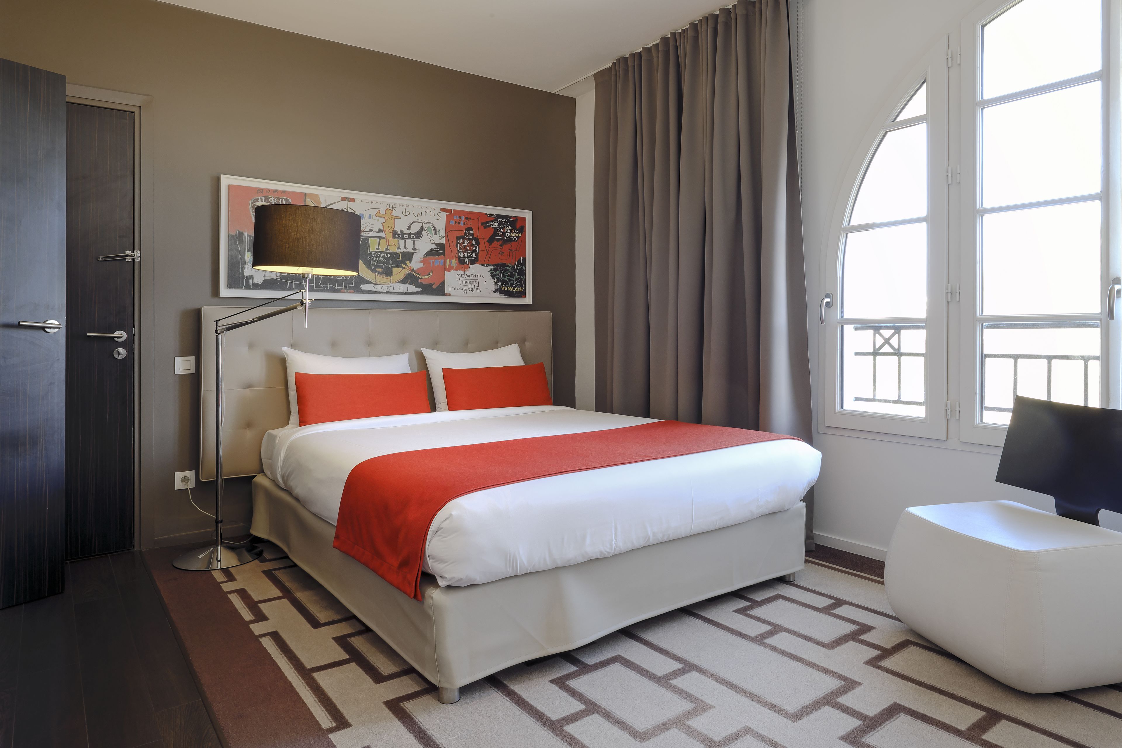 A hotel room with a large bed, a desk, a chair, a window with curtains, and a wall-mounted artwork.