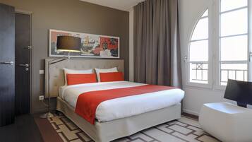 A hotel room with a large bed, a desk, a chair, a window with curtains, and a wall-mounted artwork.