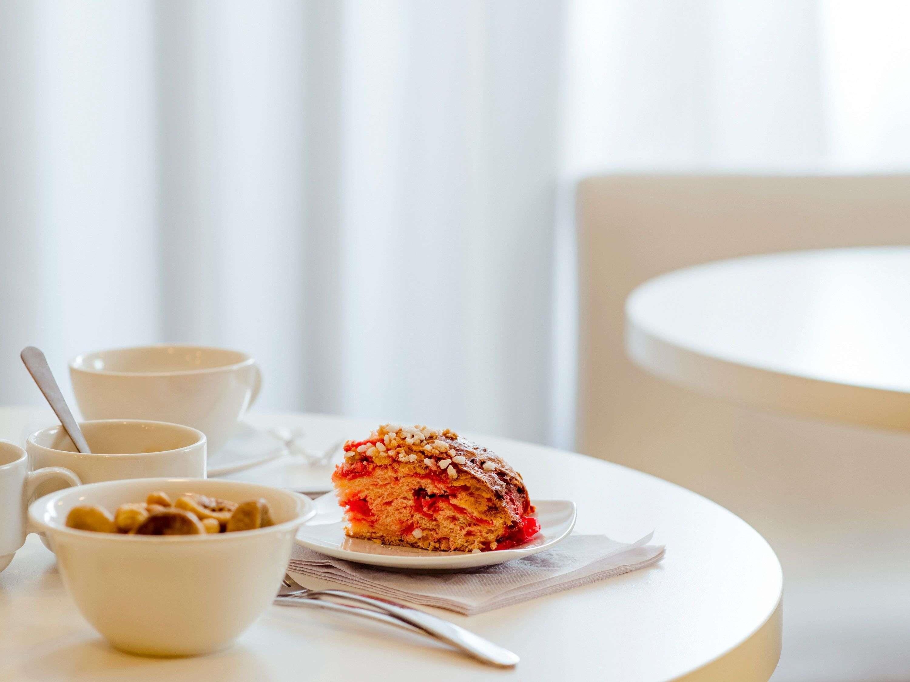 Une table avec une part de gâteau, un bol de noix et une tasse, le tout disposé dans une pièce lumineuse où se trouve une chaise blanche.