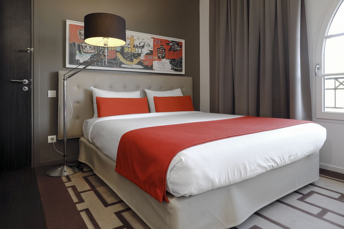 Een hotelkamer met een bed, een vloerlamp, een kunstwerk aan de muur en een raam met gordijnen.