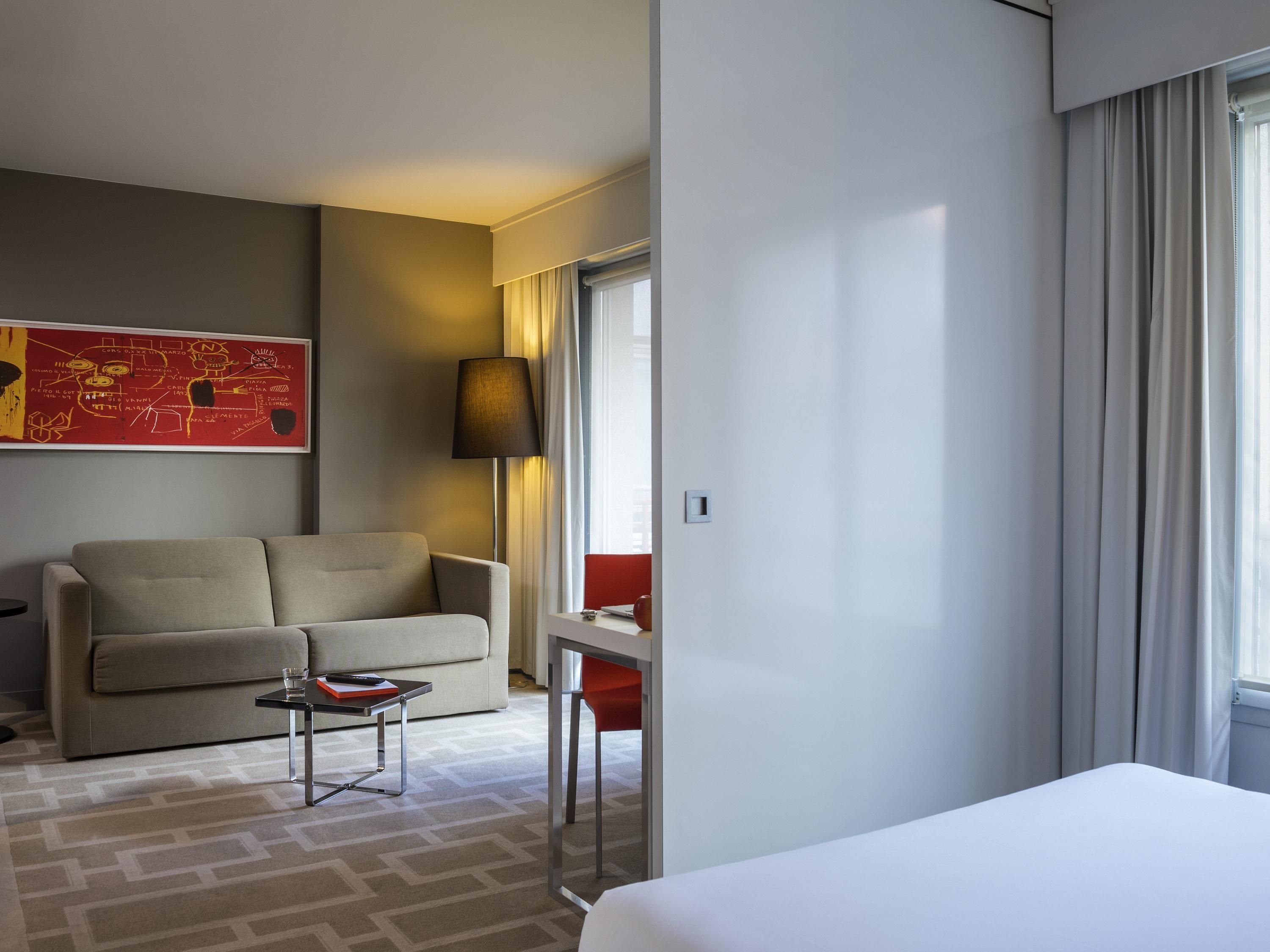 A hotel room with a bed, a sofa, a small table, a lamp, a window with curtains, and a red chair.