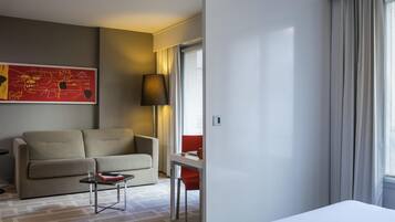 A hotel room with a bed, a sofa, a small table, a lamp, a window with curtains, and a red chair.