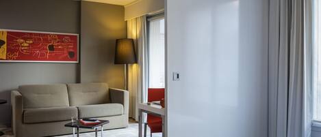 Una habitación de hotel con cama, sofá, una mesita, lámpara, ventana con cortinas y una silla roja.