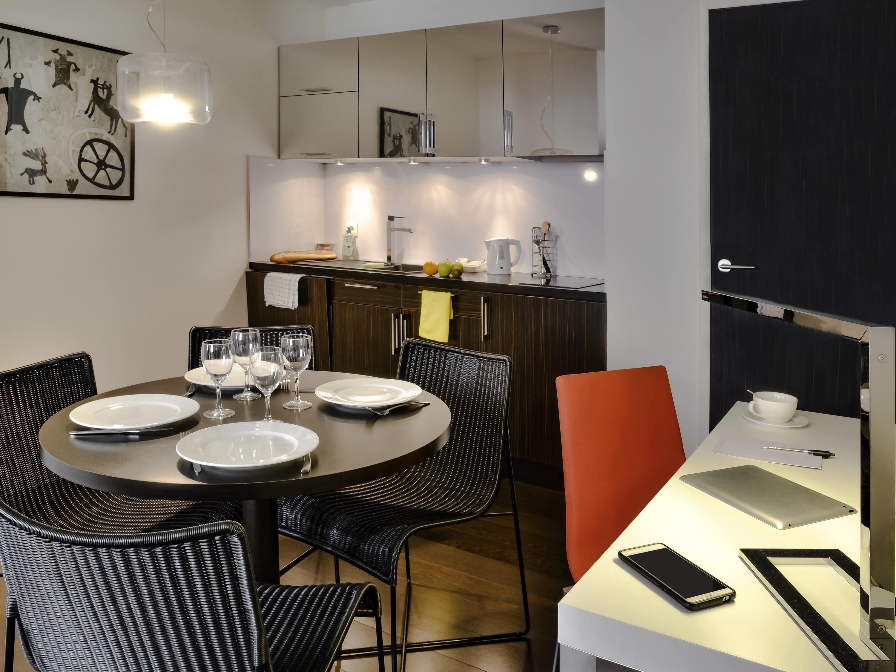 A modern dining area with a round table, black chairs, a white table, and a black chair. There is a kitchen in the background with cabinets and a countertop.