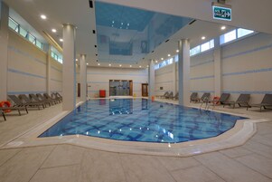 Una piscina interior con baldosas azul claro, rodeada de sillas de descanso y un techo con tragaluces.