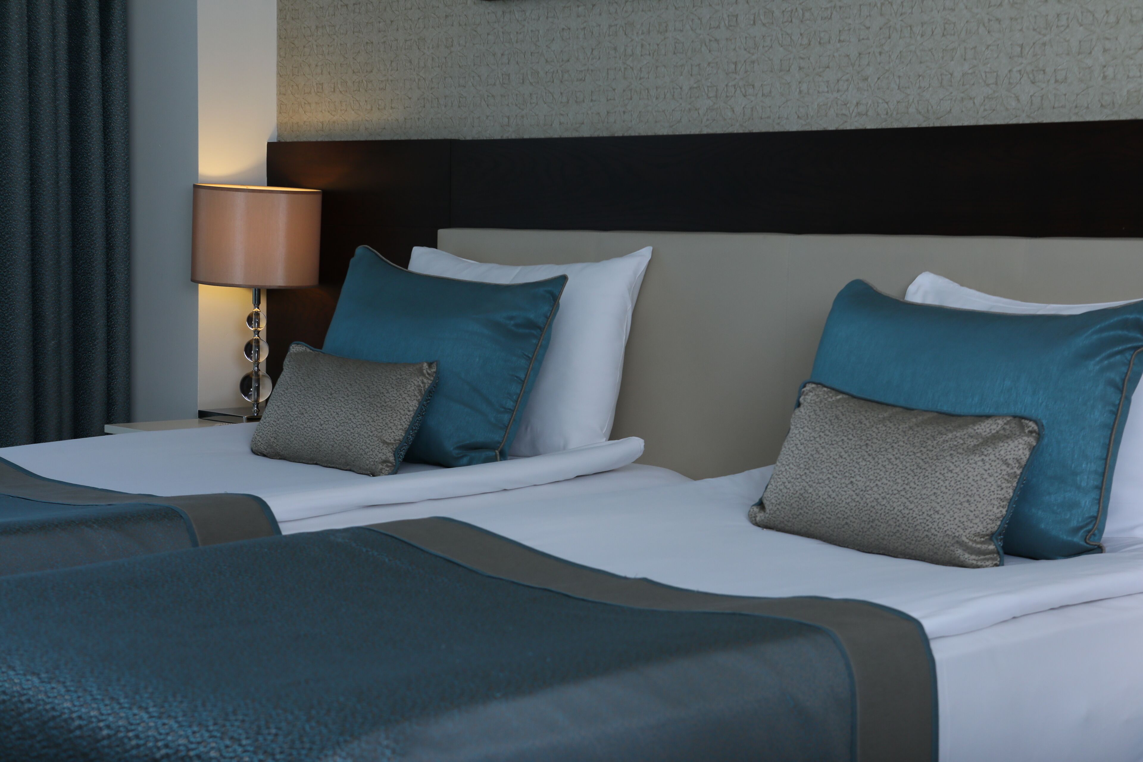 Une chambre d’hôtel avec deux lits, chacun doté d’un matelas blanc et d’oreillers turquoise et gris. Une lampe se trouve sur la table de chevet à côté des lits.