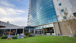 A modern hotel with a curved glass facade, a covered outdoor seating area, and a well-maintained lawn. - Baia Bursa Hotel (Bursa)