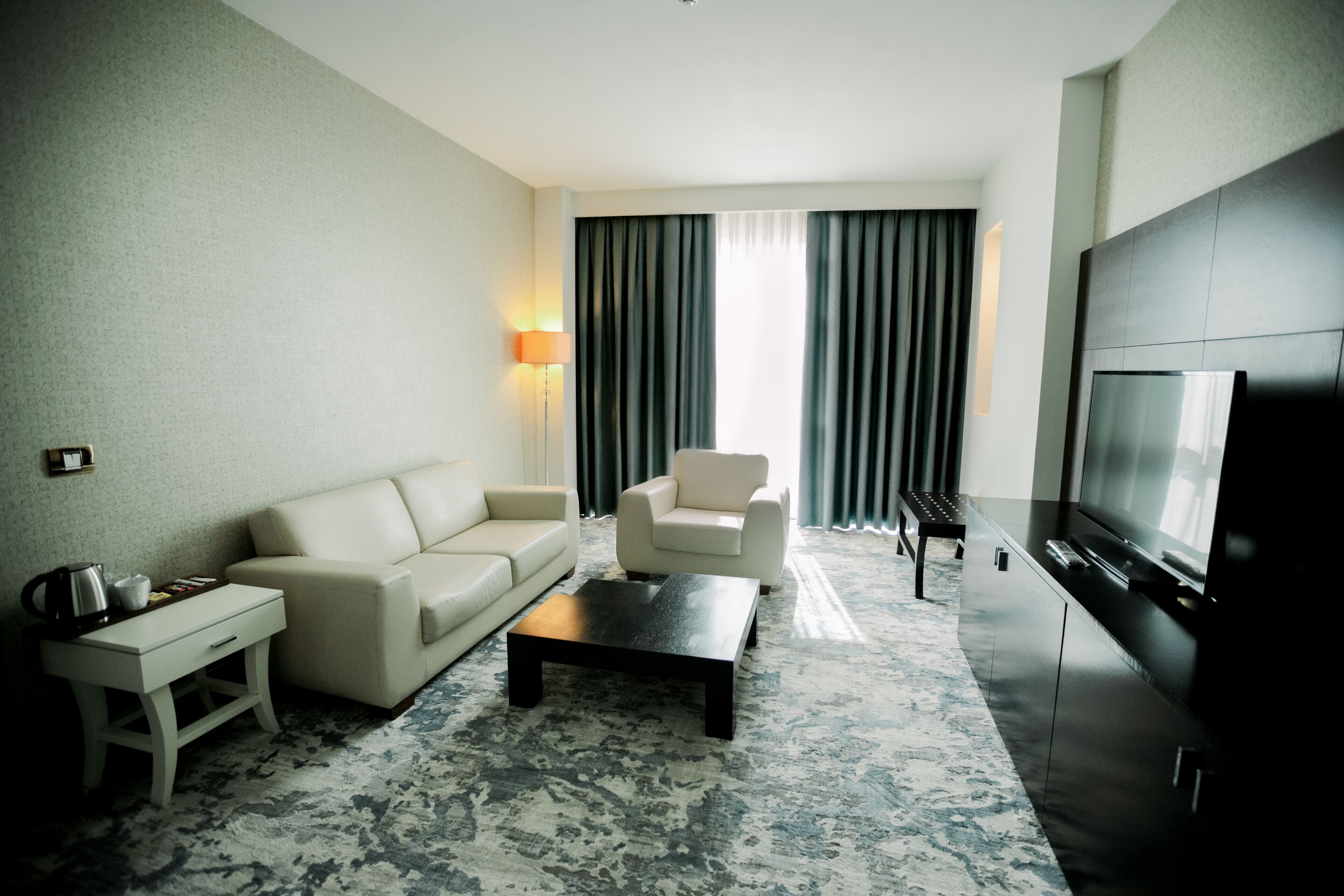 A modern hotel room with a sofa, armchair, coffee table, TV, and a lamp.