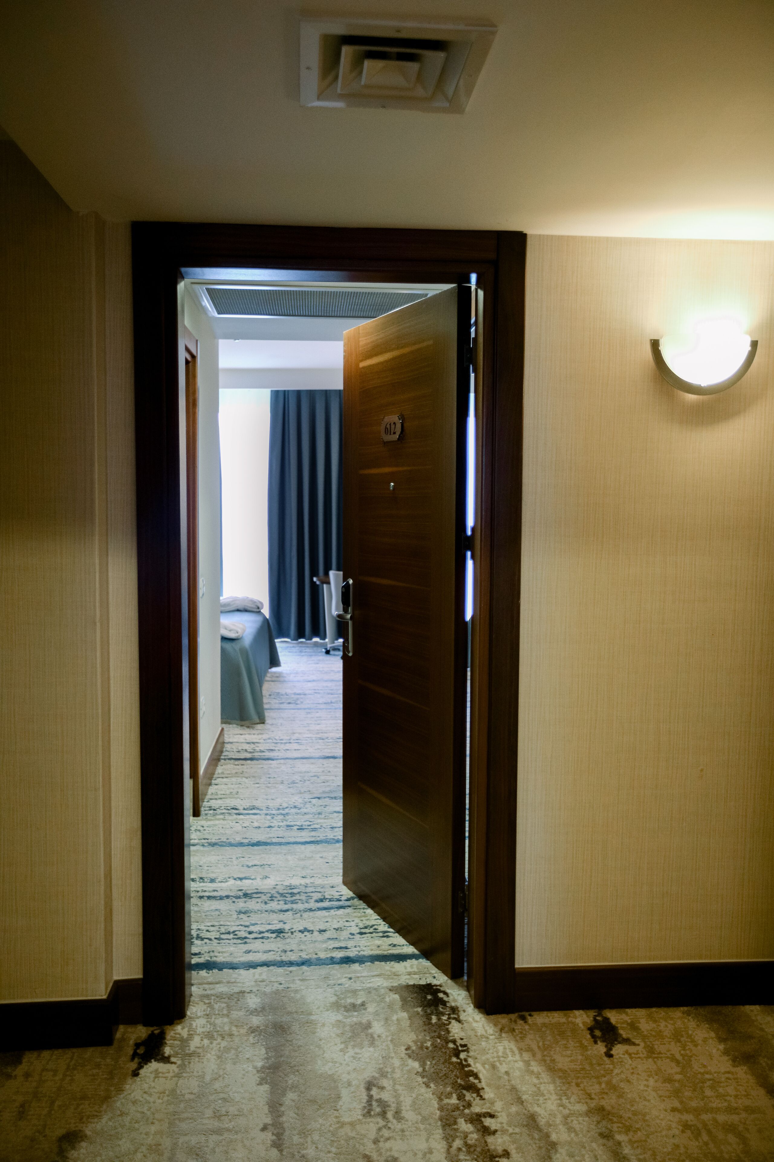 A hotel hallway leading to a room with a bed, a nightstand, and a lamp.