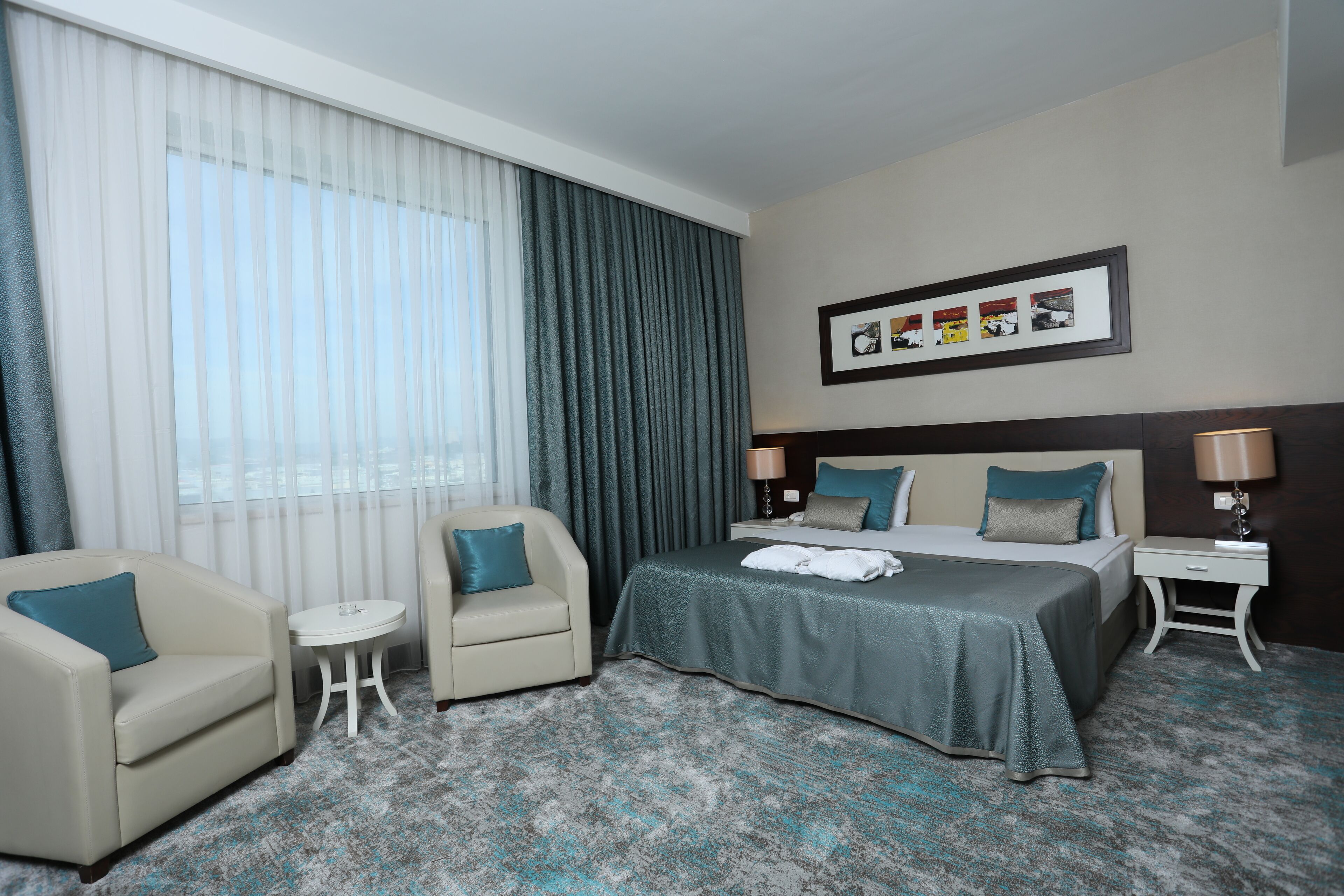 A hotel room with a bed, two chairs, a small table, a nightstand with a lamp, and a framed photo on the wall.