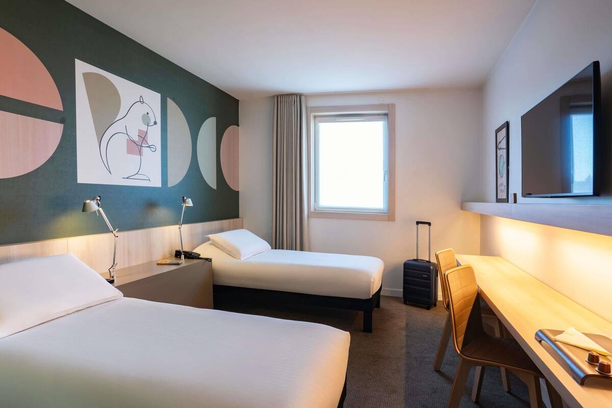 Une chambre d’hôtel avec deux lits, un bureau, une chaise, une lampe de bureau, une valise, une fenêtre avec des rideaux et un mur décoré d’œuvres d’art abstraites.