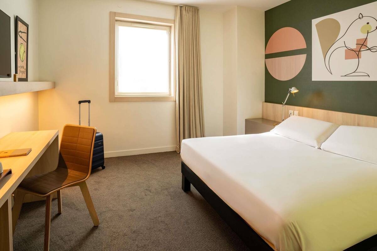 Une chambre d’hôtel avec un lit, un bureau, une chaise, une valise, une fenêtre et un mur orné d’œuvres d’art abstraites.