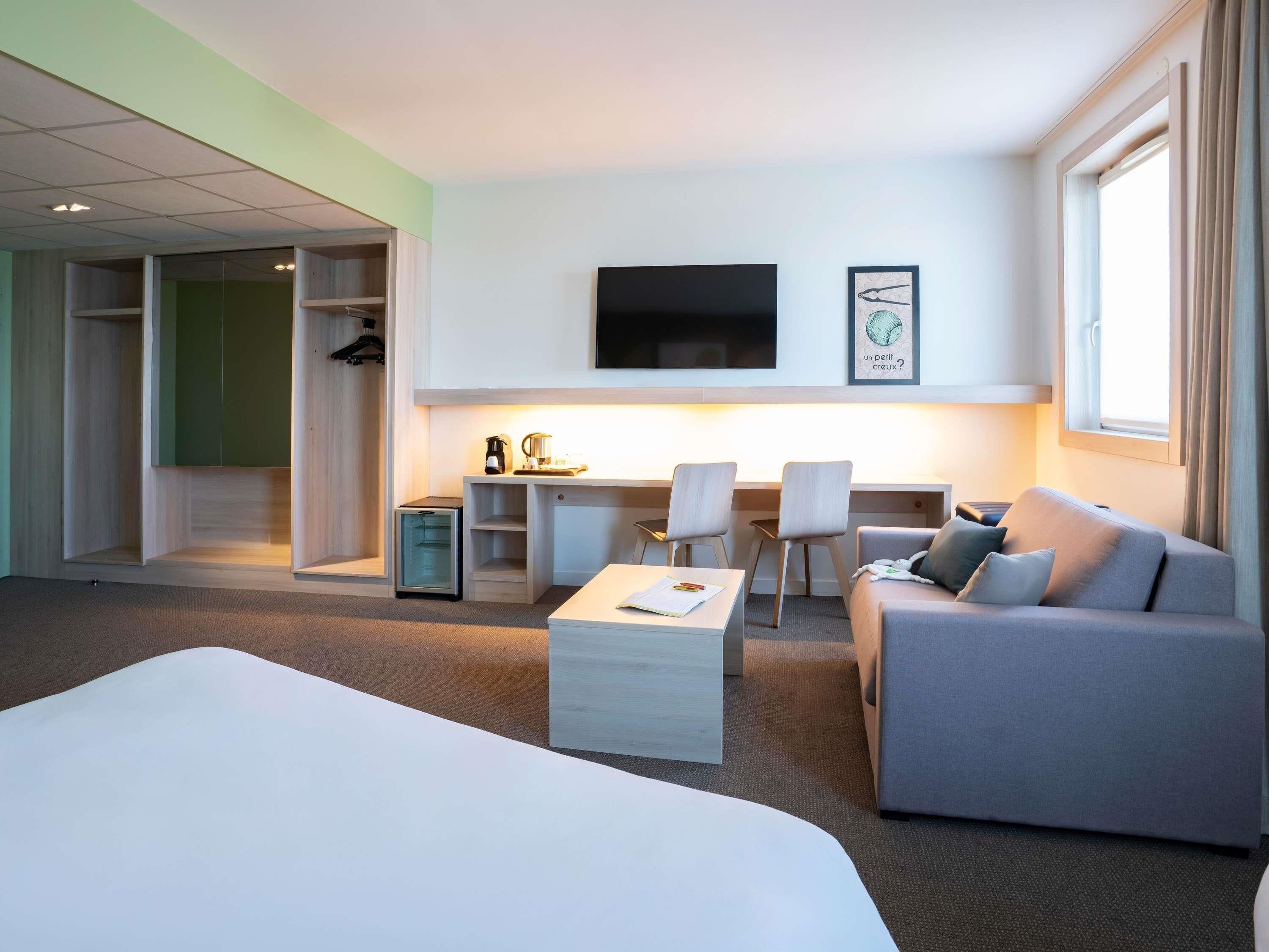 Quarto de hotel com cama, sofá, escrivaninha com cadeiras, televisão e uma pequena cozinha.