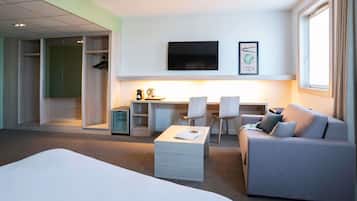 Quarto de hotel com cama, sofá, escrivaninha com cadeiras, televisão e uma pequena cozinha.