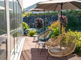 Une terrasse extérieure avec des chaises et une table en osier, une voiture garée à proximité et un mur de pierre en arrière-plan.