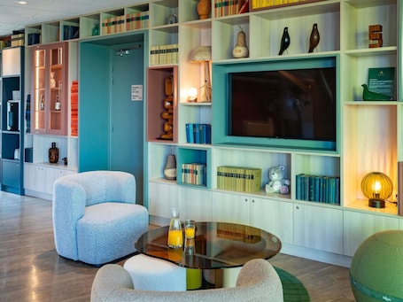 A modern living room with a glass coffee table, comfortable seating, a flat-screen TV, and a bookshelf filled with books and decorative items.. ibis Styles Guéret