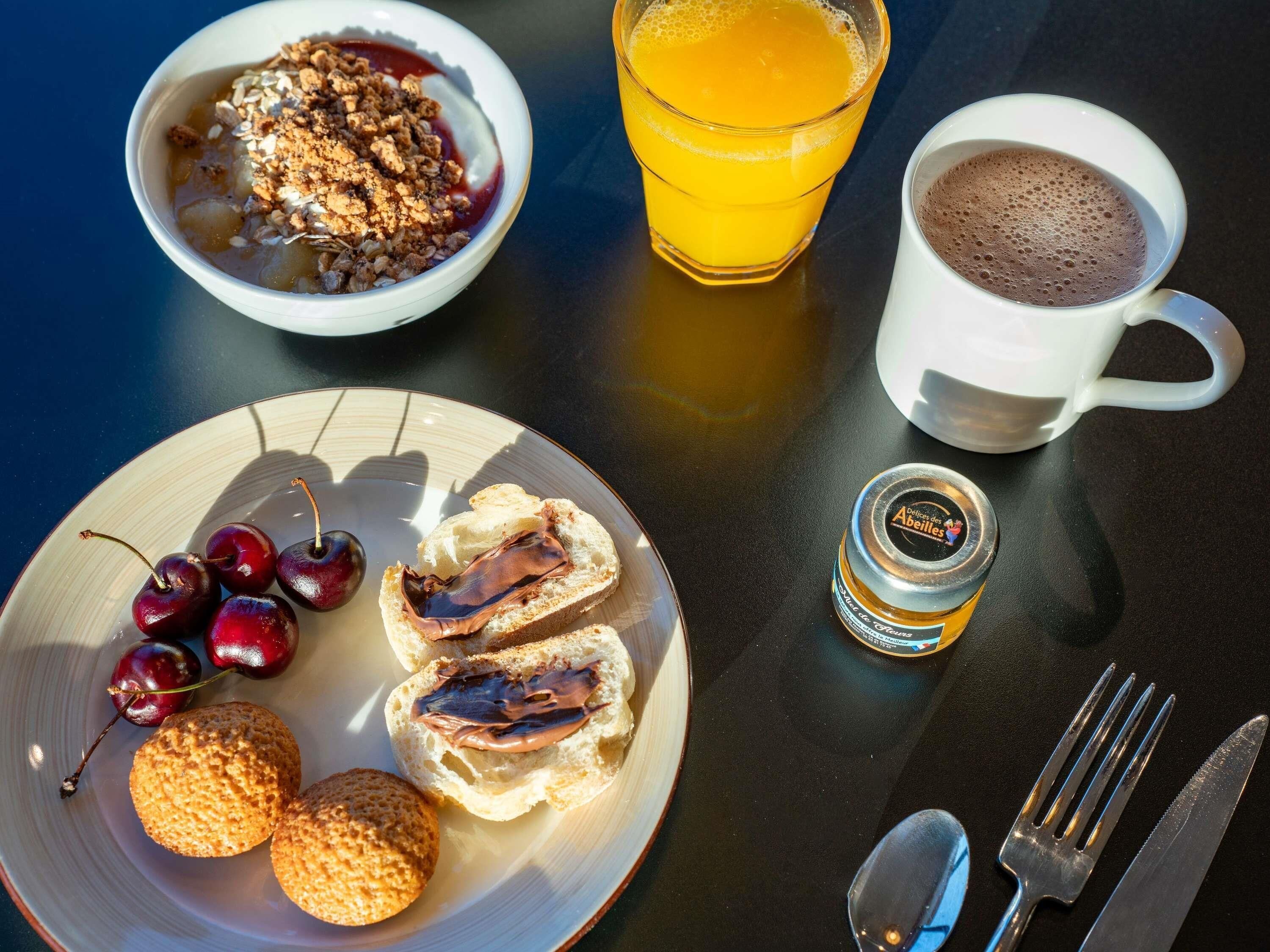 A breakfast table with a bowl of cereal, a glass of orange juice, a cup of hot chocolate, a plate with biscuits and cherries, and a jar of jam.
