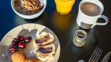 A breakfast table with a bowl of cereal, a glass of orange juice, a cup of hot chocolate, a plate with biscuits and cherries, and a jar of jam.