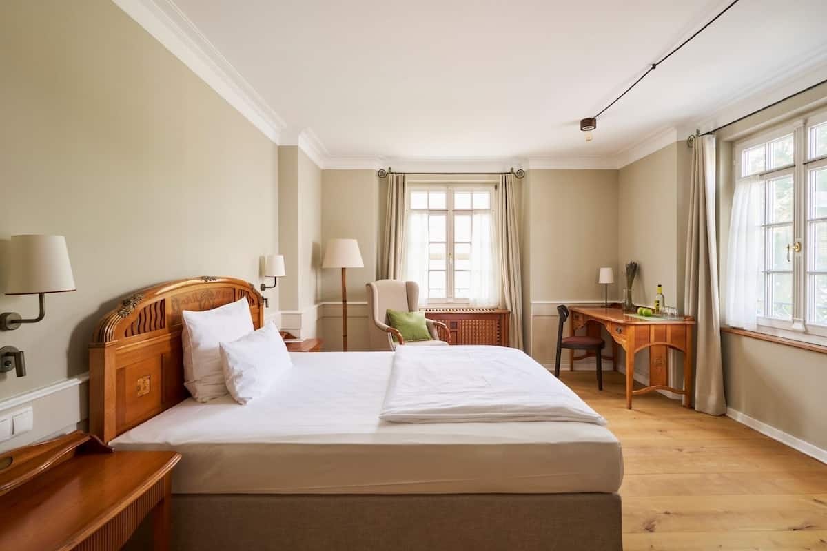 A hotel room with a large bed, a desk, a chair, and a window with curtains.