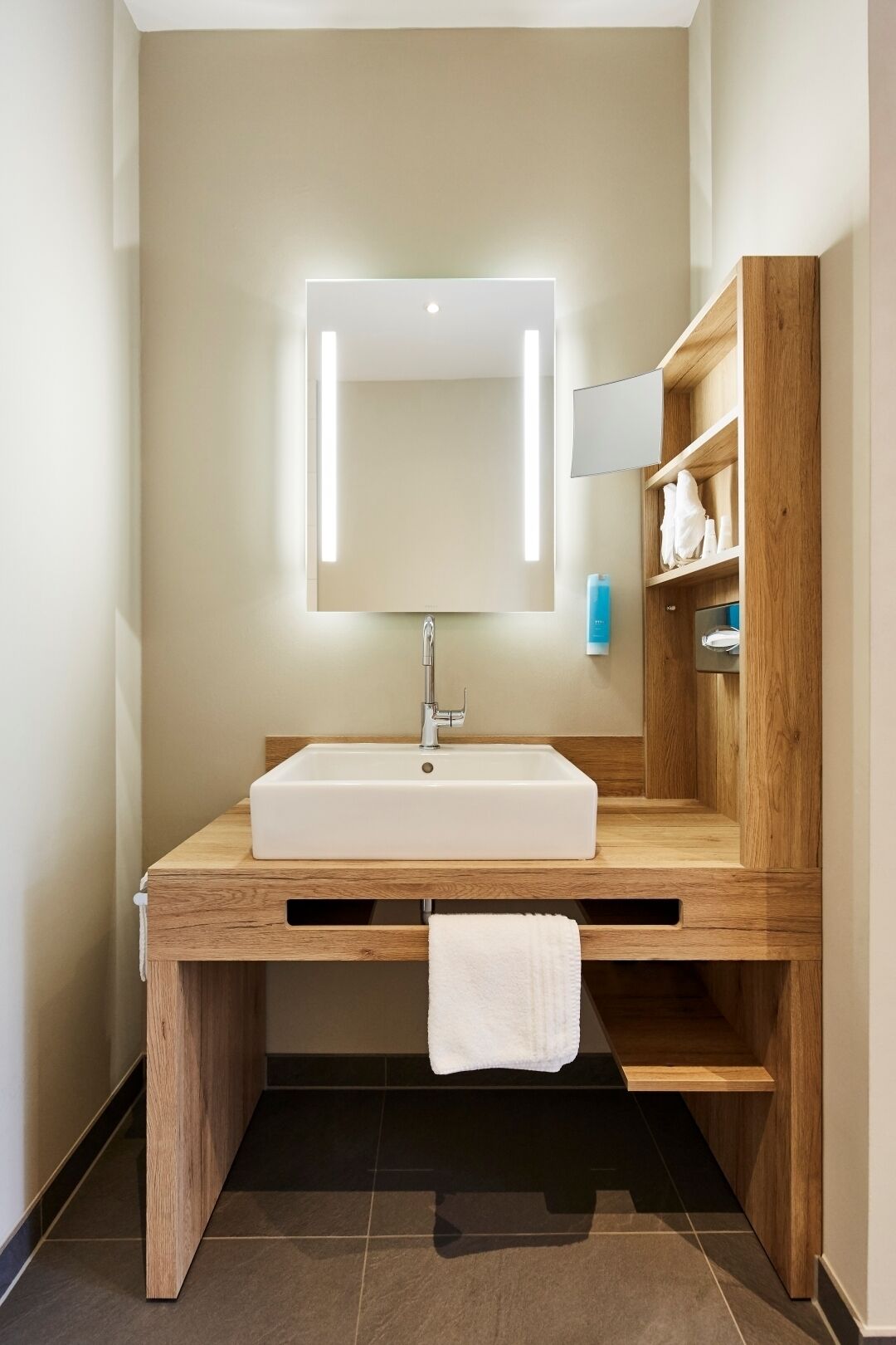 Une salle de bain moderne avec un lavabo rectangulaire, un miroir doté d’un éclairage intégré, des meubles en bois et un porte-serviettes.