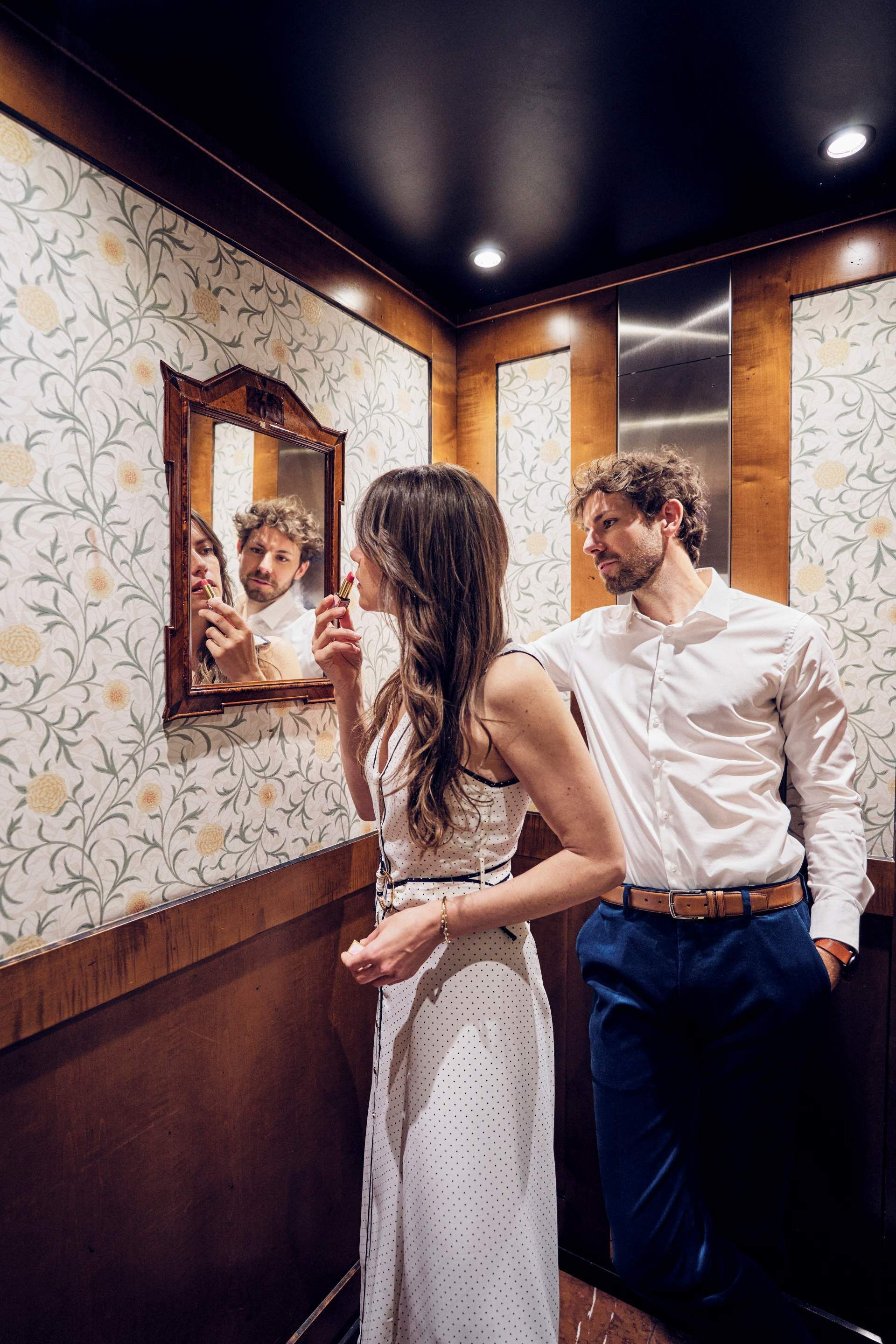 A woman applying lipstick in front of a mirror in a room with floral wallpaper and wooden paneling.