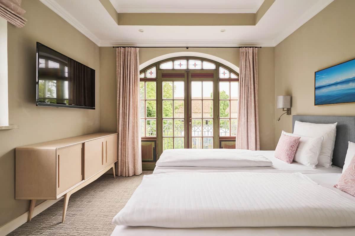 A hotel room with a large bed, a TV mounted on the wall, a view of greenery through the window, and a painting above the bed.