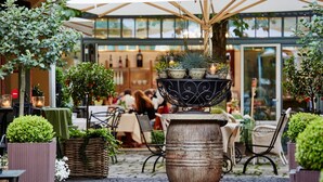 Un espace de café en plein air avec des tables et des chaises, un grand bac à fleurs décoratif au centre, et une vitrine en verre en arrière-plan.