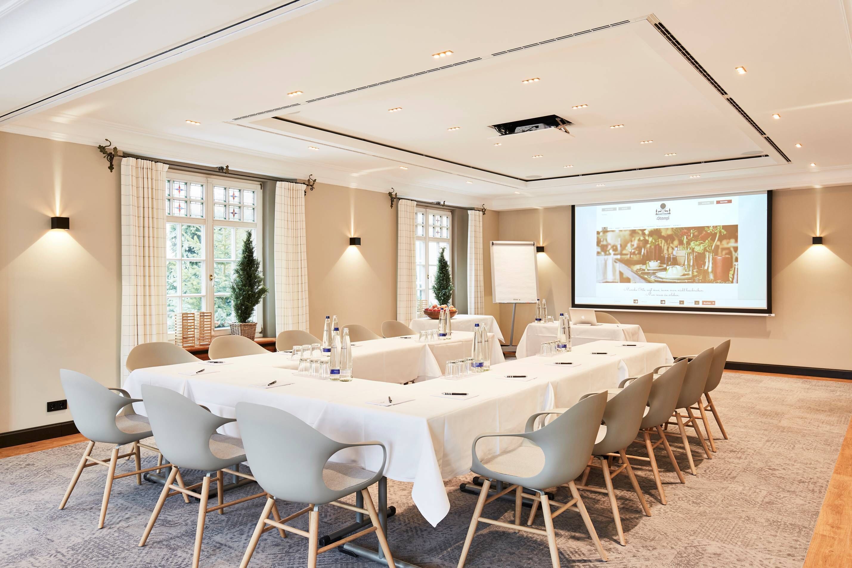 Ein Konferenzraum mit einem langen Tisch, Stühlen, einer Projektionsleinwand und einem großen Fenster mit Vorhängen.