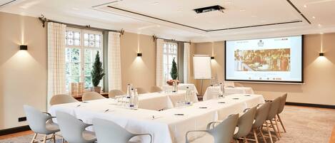 Un salón de conferencias con una larga mesa, sillas, una pantalla de proyector y un amplio ventanal con cortinas.