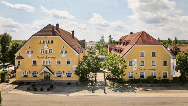 Un bâtiment jaune portant les enseignes « Gasthof Stangl » et « Hôtel Stangl ». Il présente un style architectural européen traditionnel, avec un toit à forte pente et de nombreuses fenêtres.