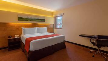 A hotel room with a bed, a desk, a chair, a nightstand, and a window with a view.