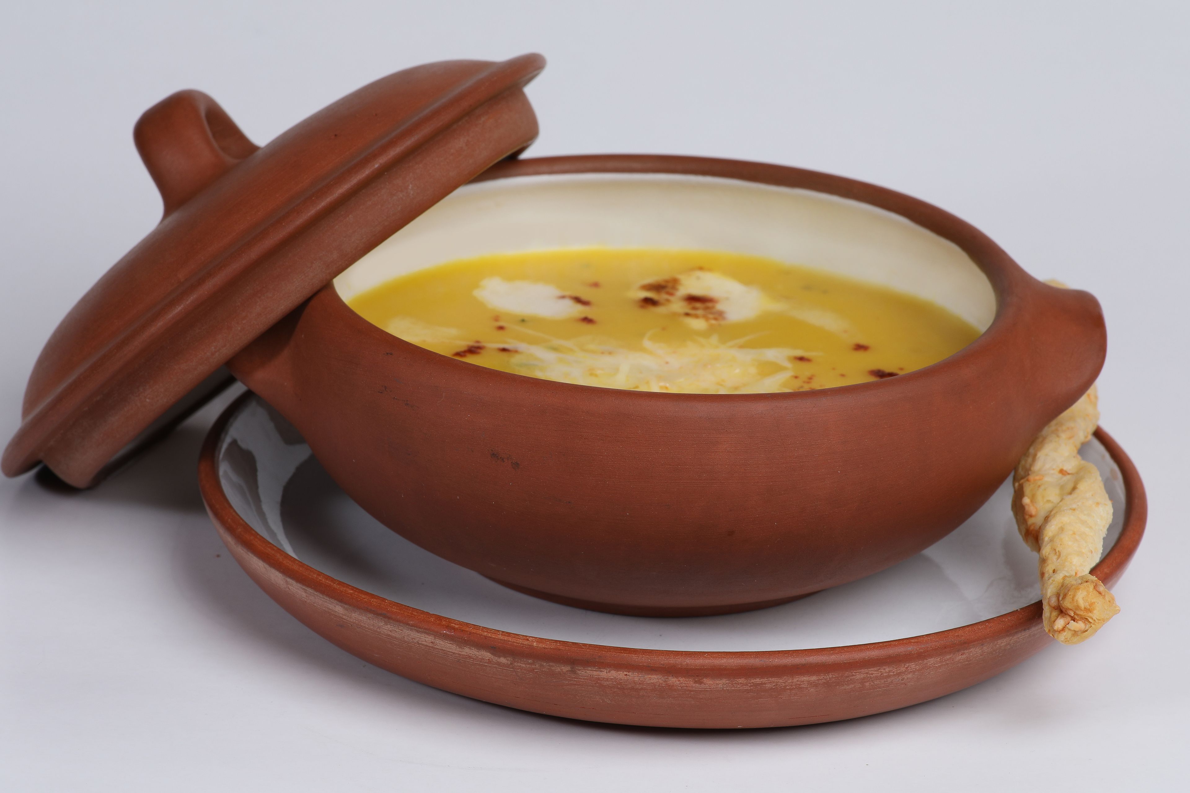 Una olla de barro con tapa que contiene una sopa amarilla con ingredientes visibles.
