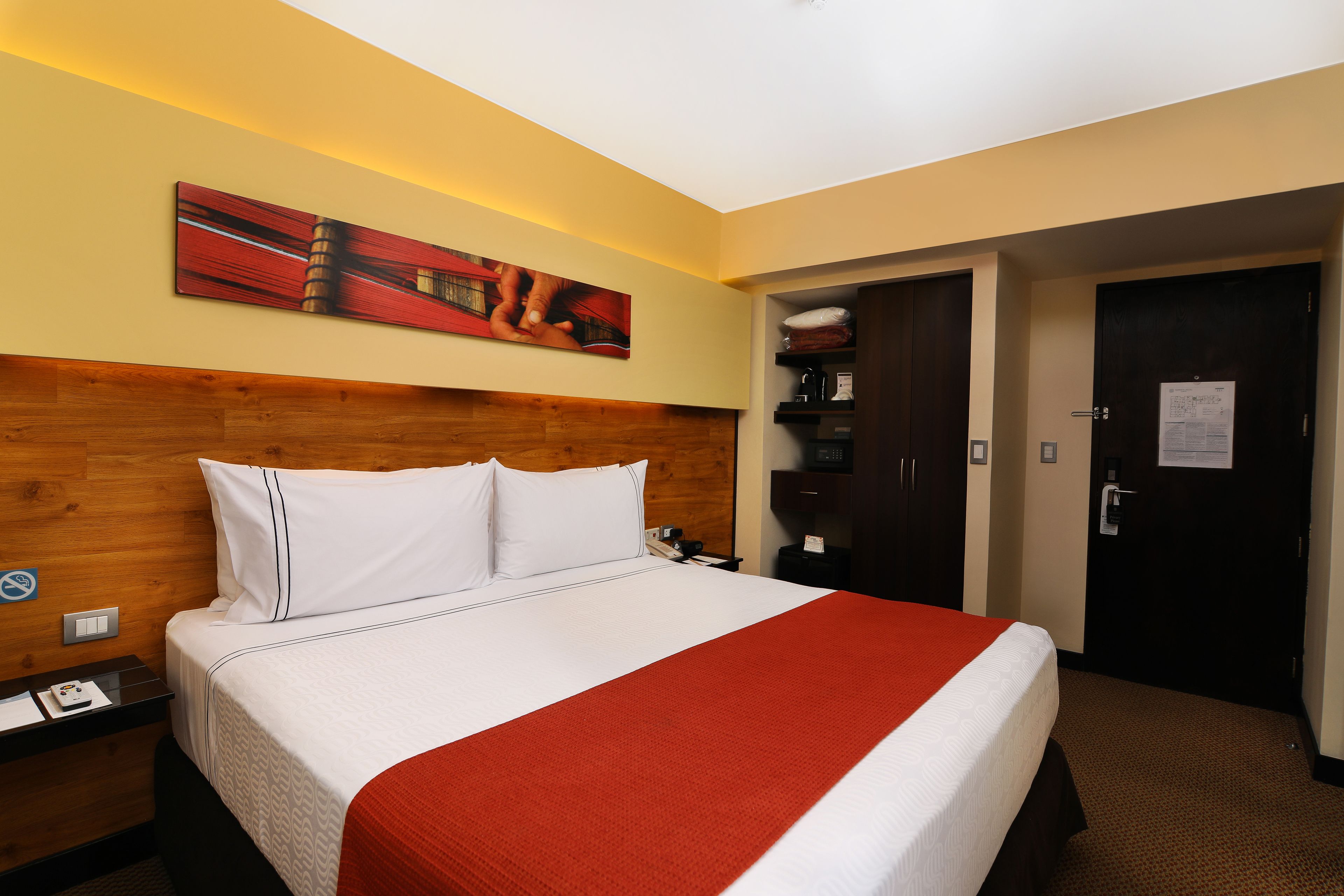 Habitación de hotel con cabecera de madera, una cama grande con ropa de cama blanca y roja, un cuadro en la pared y una puerta con un letrero.