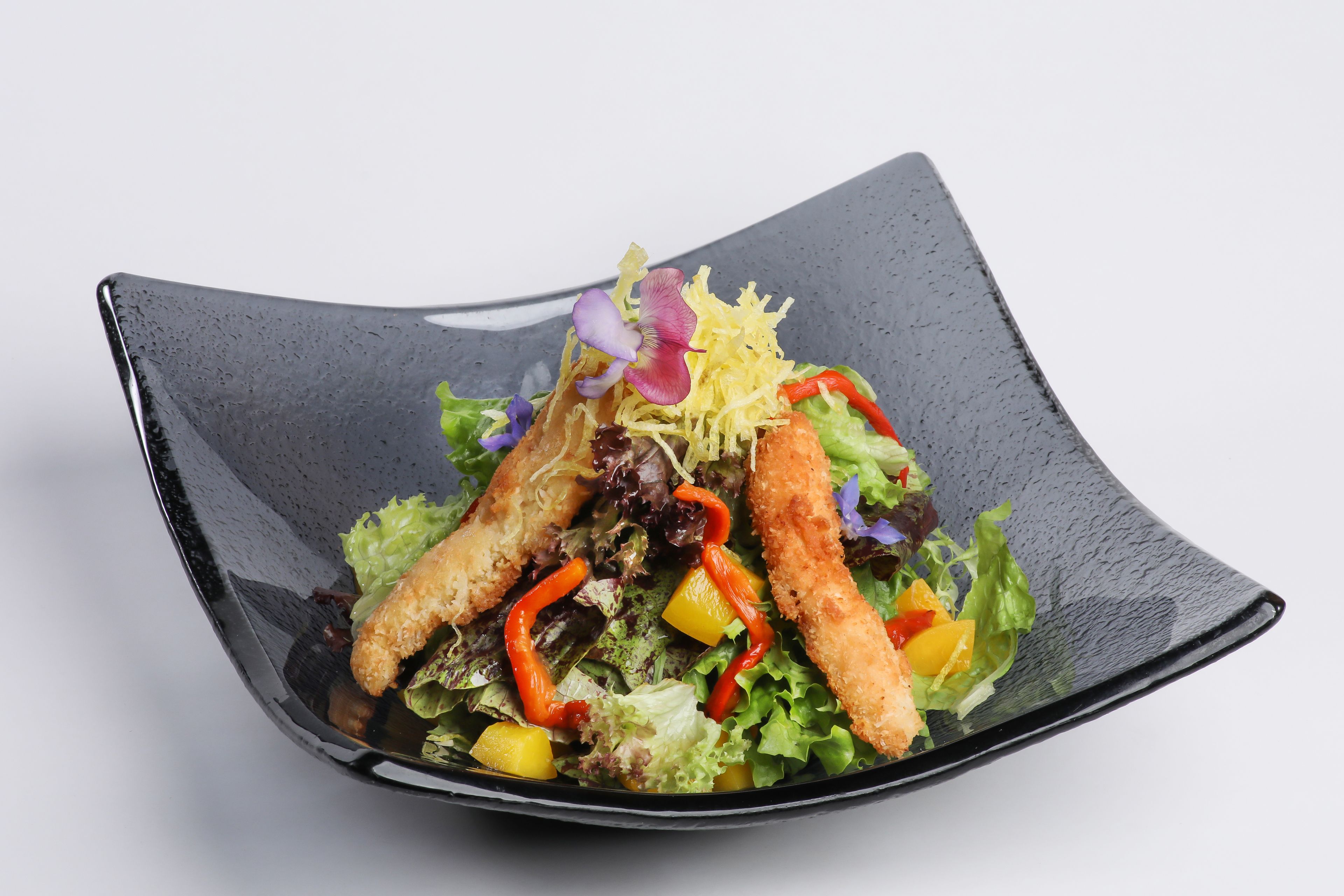 A square black plate with a salad containing lettuce, red peppers, yellow bell peppers, and a purple flower.