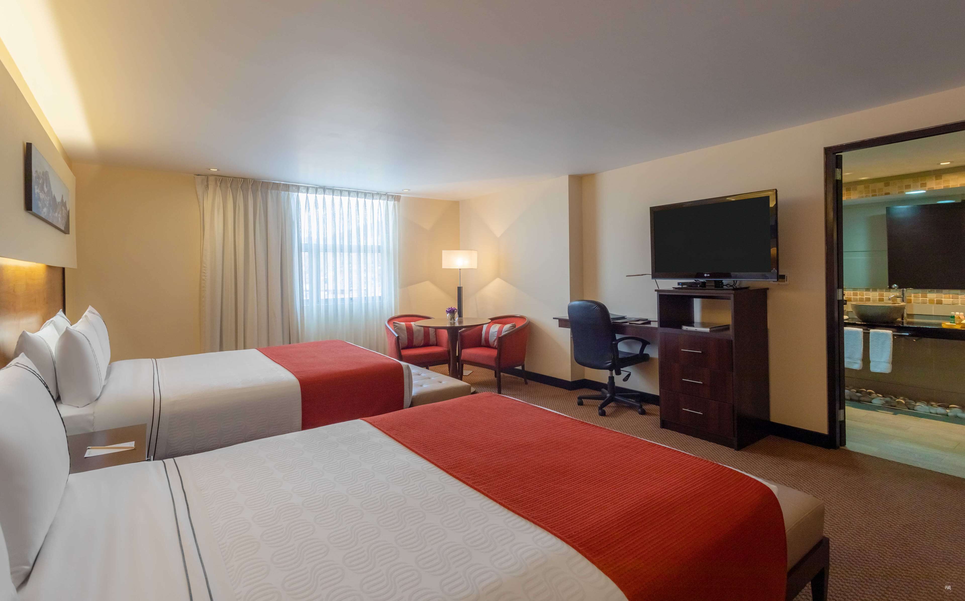 Une chambre d’hôtel avec deux lits, un bureau, une chaise, une télévision et une salle de bain visible à travers une porte en verre.