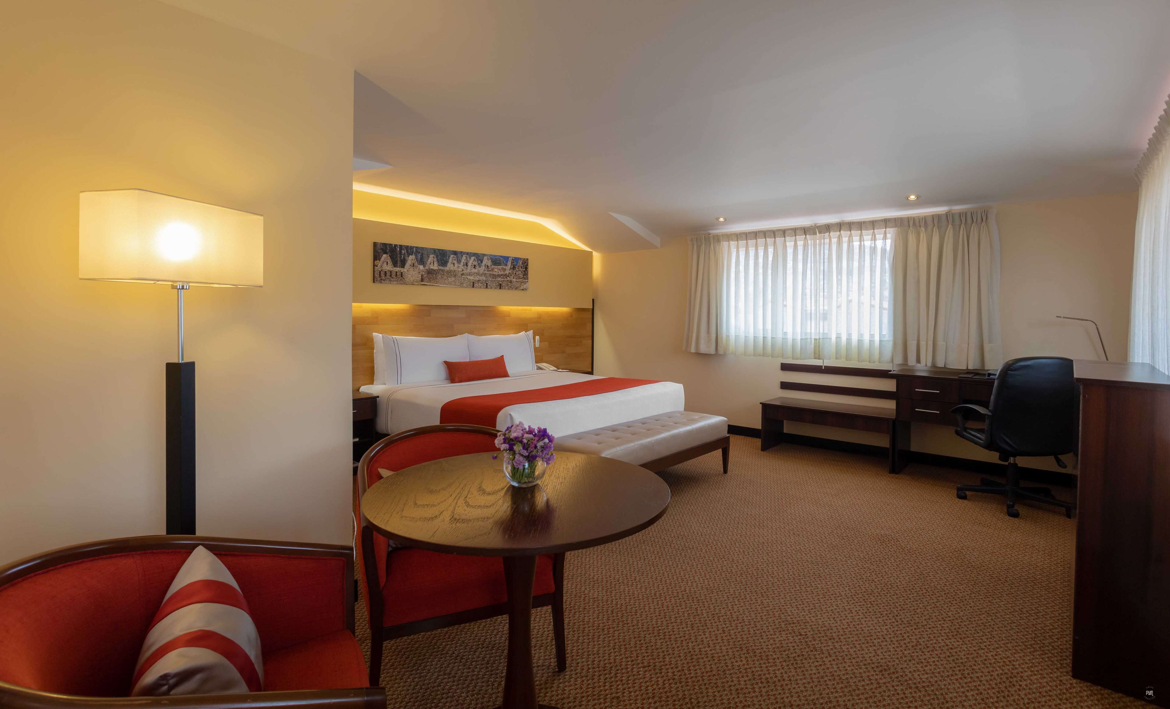 A hotel room with a bed, bedside table, lamp, desk, chair, and a small table with a vase of flowers.