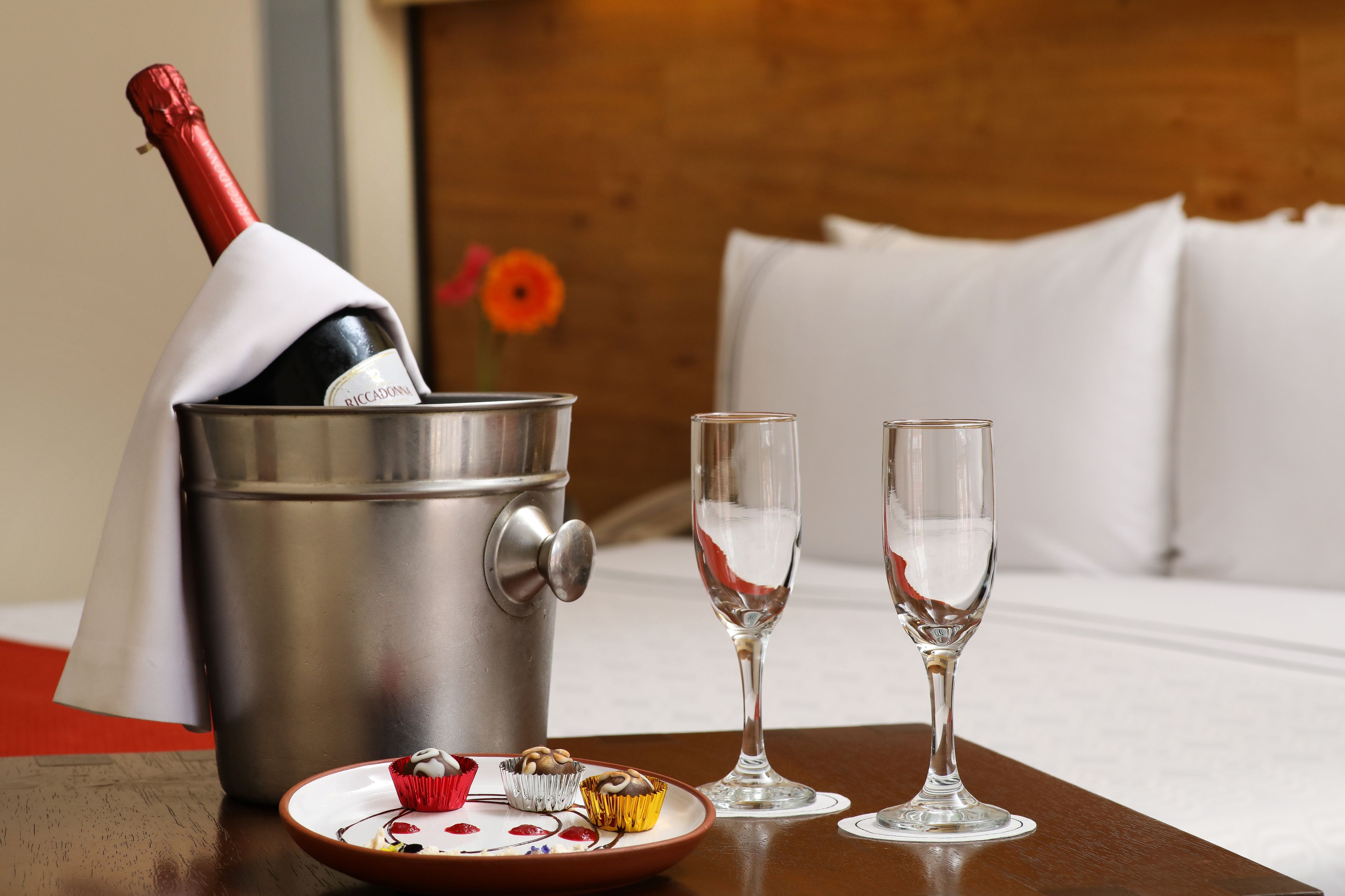 A champagne bottle in a bucket, two glasses, and a plate with cupcakes and a chocolate bar on a wooden table in a hotel room.