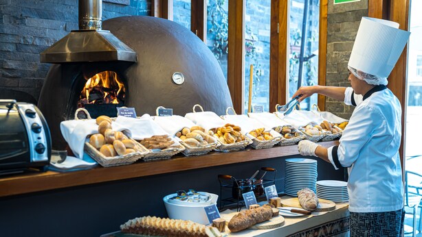 A chef preparing food in a kitchen with a wood-fired oven, various breads, and pastries on display. - Sonesta Hotel Cusco (Cusco)