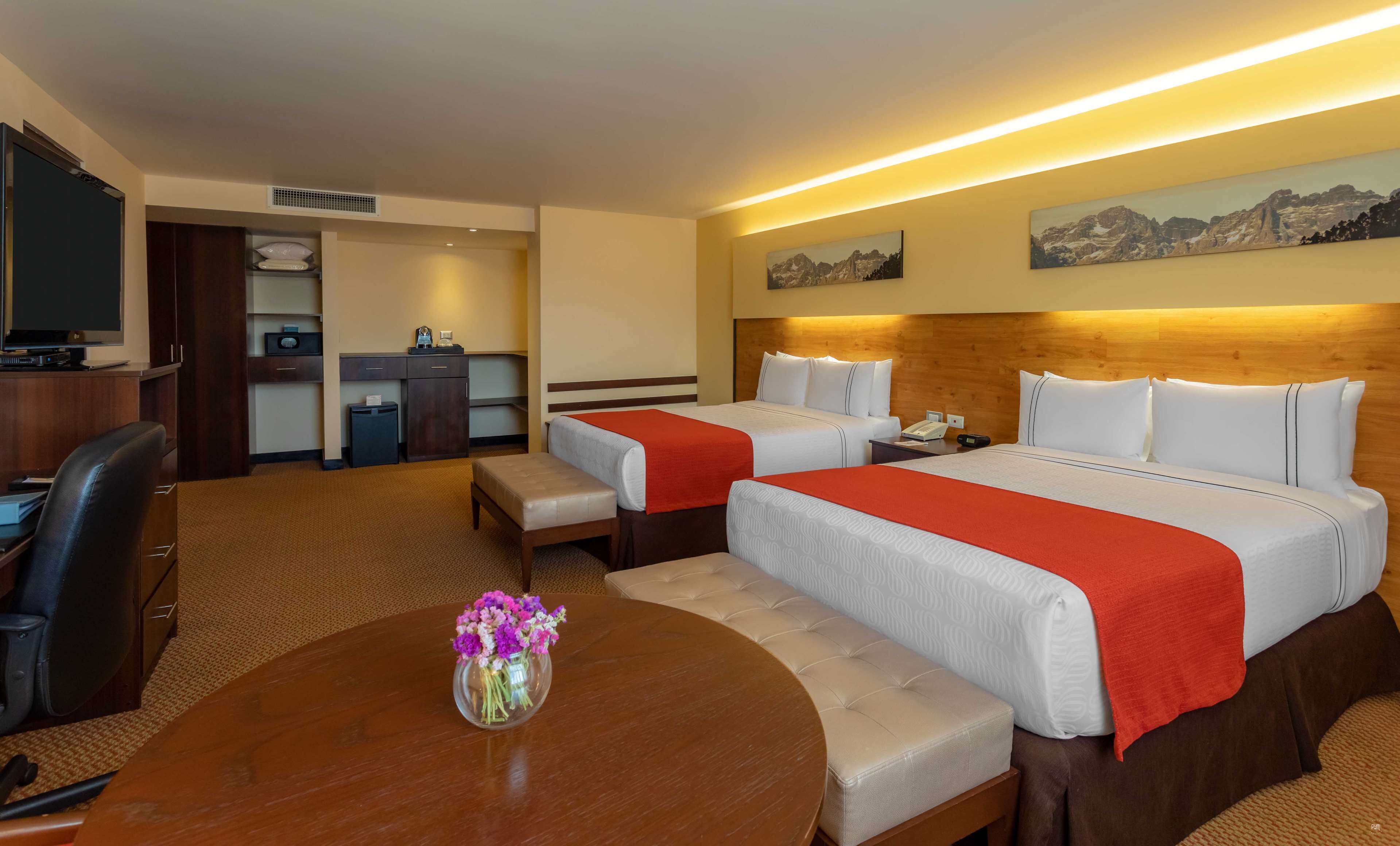 A hotel room with two beds, a desk, a TV, a small kitchenette, and a dining table with a vase of flowers.