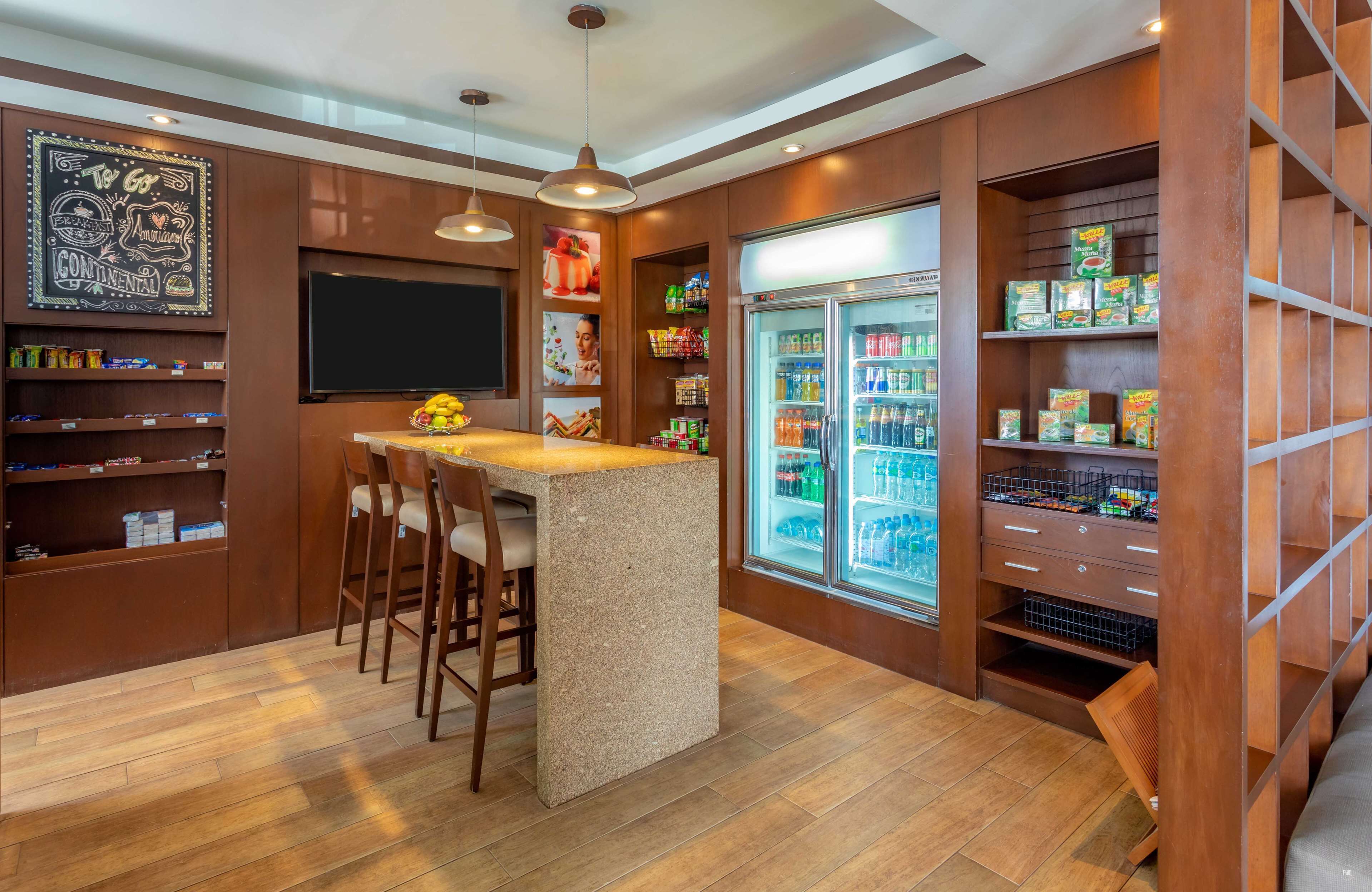 A modern bar area with a central island, bar stools, a refrigerator stocked with beverages, and shelves with snacks.