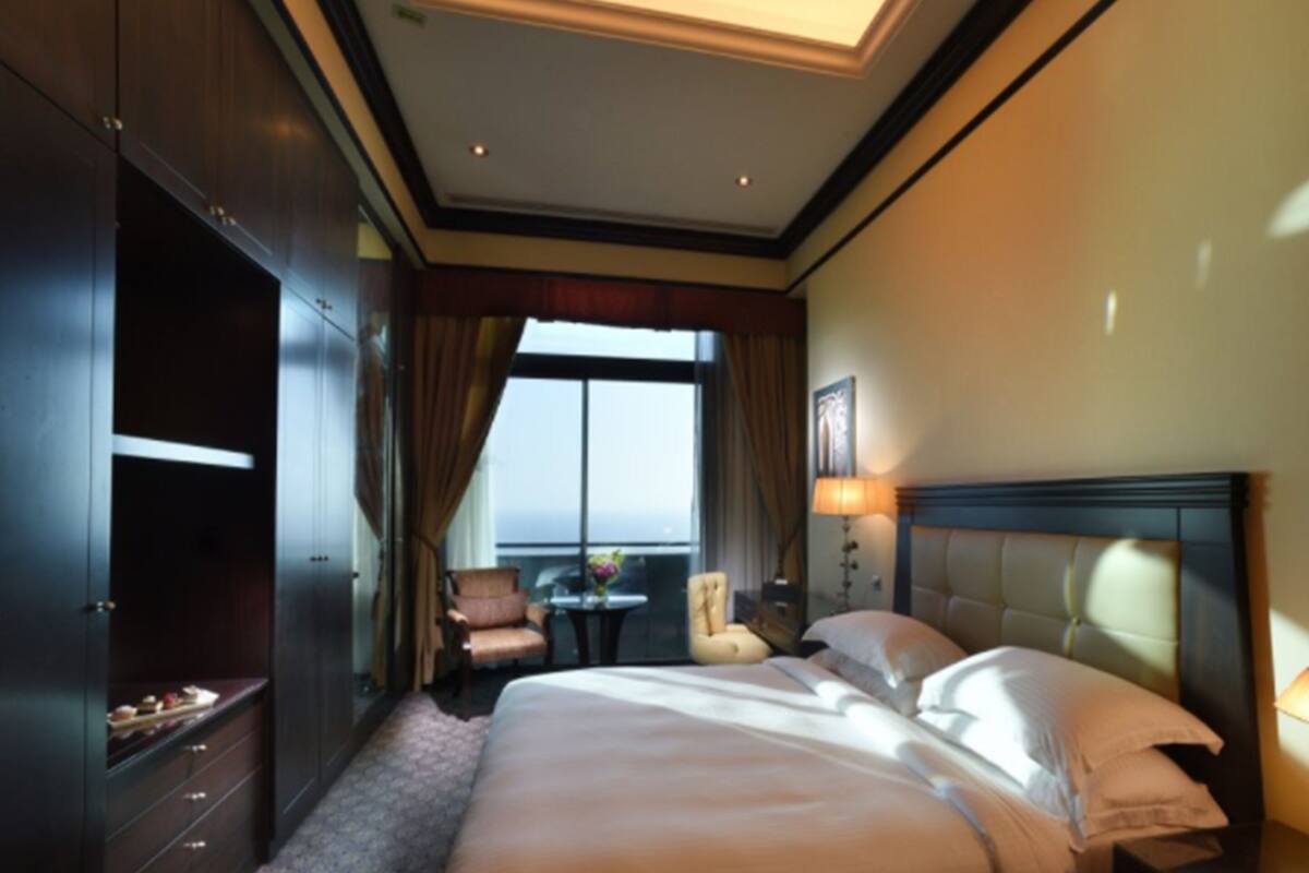 Habitación de hotel con una cama grande, un armario, un área de estar con una mesa y sillas, y vistas al exterior a través de una ventana.