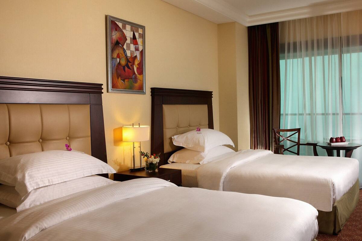 Habitación de hotel con dos camas, una mesita de noche con lámpara, un cuadro en la pared y vistas al exterior a través de una ventana con cortinas.