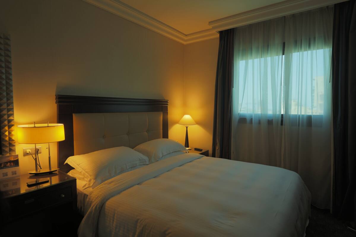 Una habitación de hotel con una cama grande, dos lámparas de noche y un ventanal con cortinas traslúcidas.